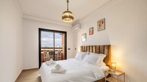 A modern bedroom with a large bed, a wooden headboard, a bedside table with a lamp, and a view through a glass door to the outside. - Guéliz 2BR Near M Avenue – Piscine, Balcons & Clim (Marrakesh)
