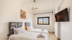A hotel room with two beds, a mounted TV displaying the Netflix logo, a painting on the wall, and a window with a city view. - Guéliz 2BR Near M Avenue – Piscine, Balcons & Clim (Marrakesh)