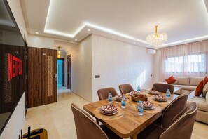 A modern dining area with a wooden table set for four, surrounded by chairs. There is a chandelier above the table, and the room has a neutral color palette with beige walls and a large window with curtains. - 2 min M-avenue / Guéliz Clim, Piscines & Parking (Marrakesh)