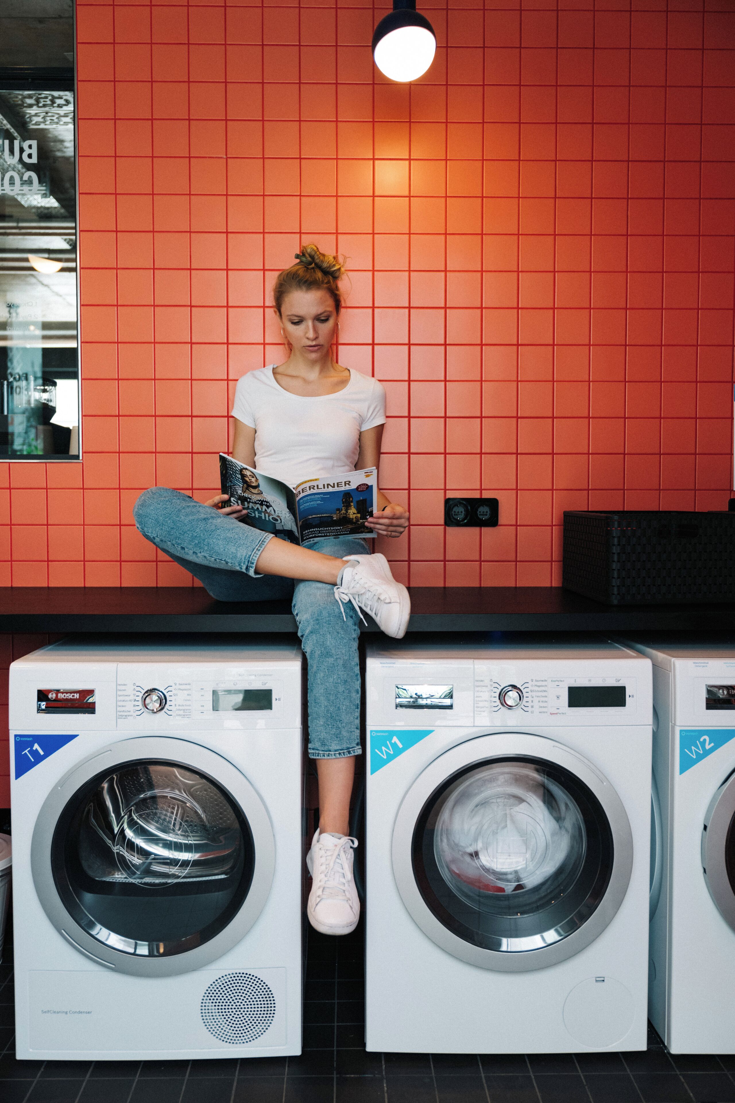 A person sitting on top of two stacked washing machines in a room with red tiled walls.