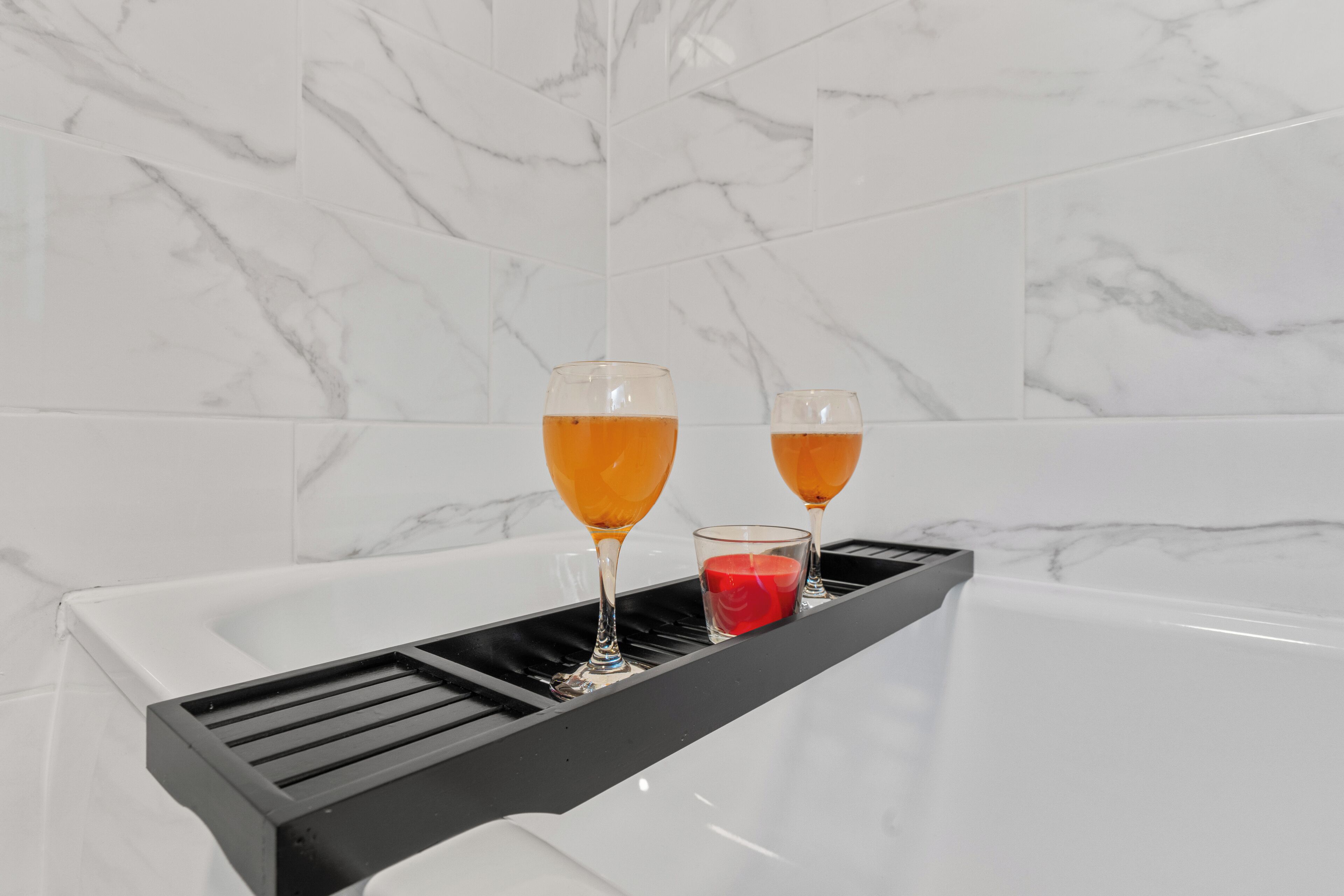 A bathroom with a marble wall and a black tray holding two glasses of amber liquid and a red candle.
