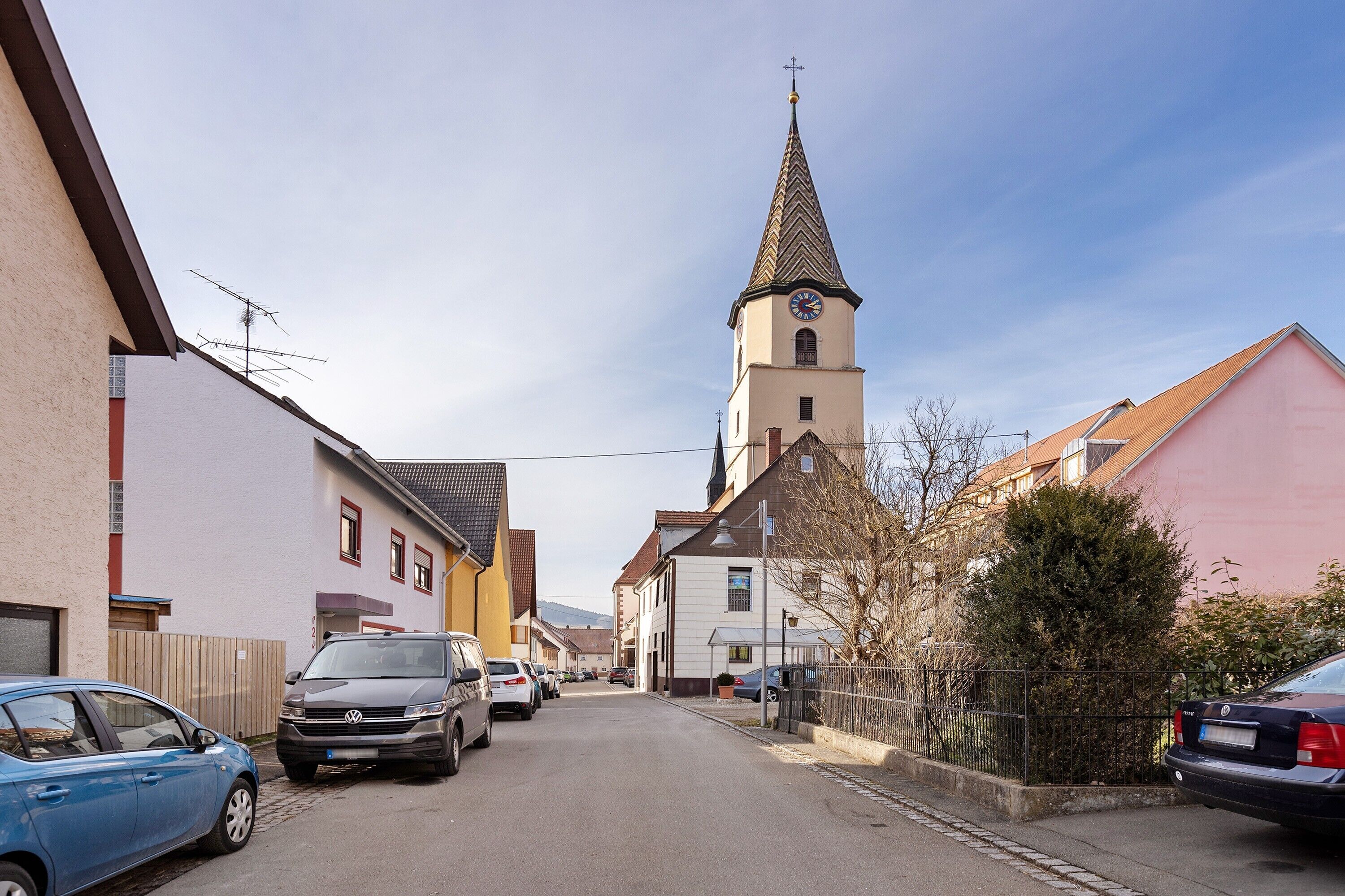 A street with parked cars, a church with a tall steeple, and residential buildings.