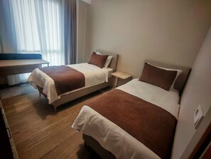 Hotellihuone, jossa on kaksi sänkyä, puulattia, pieni pöytä ja ikkuna, jossa on verhot.
