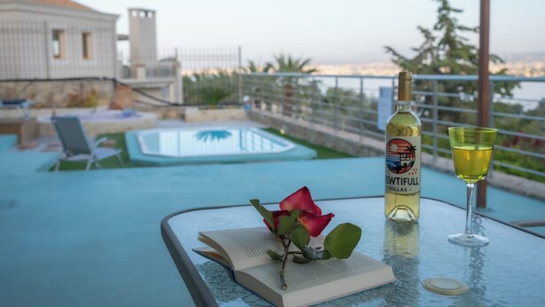 A glass table with a book, a bottle, and a glass of wine. - Viewtifull-Villas 2 – Private Pool, Playground & Panoramic Views Near Heraklion (CRETE)