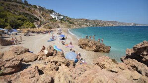 A beach with rocky terrain, clear blue water, and white buildings on a hillside. - Viewtifull-Villas 2 – Private Pool, Playground & Panoramic Views Near Heraklion (CRETE)