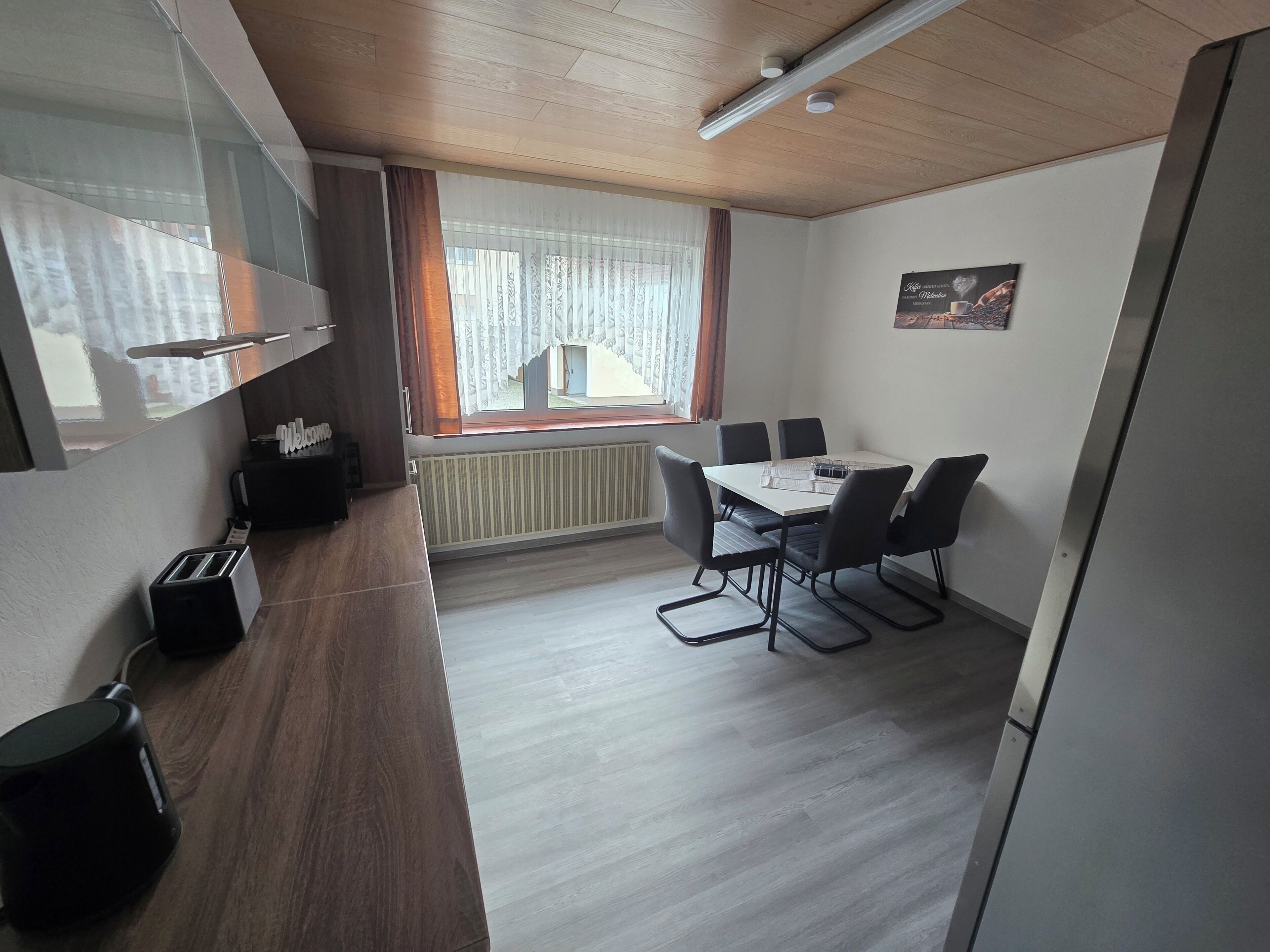 A modern room with a wooden ceiling, a dining table with four chairs, a radiator, and a window with curtains.