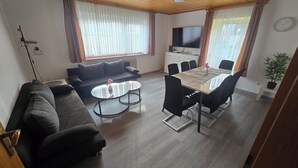 A modern living room with a sofa, a dining table with chairs, a flat-screen TV, and a potted plant. - Vacation home Lautertal-Lodge with private pool/whirlpool for up to 10 people (Kreimbach-Kaulbach)
