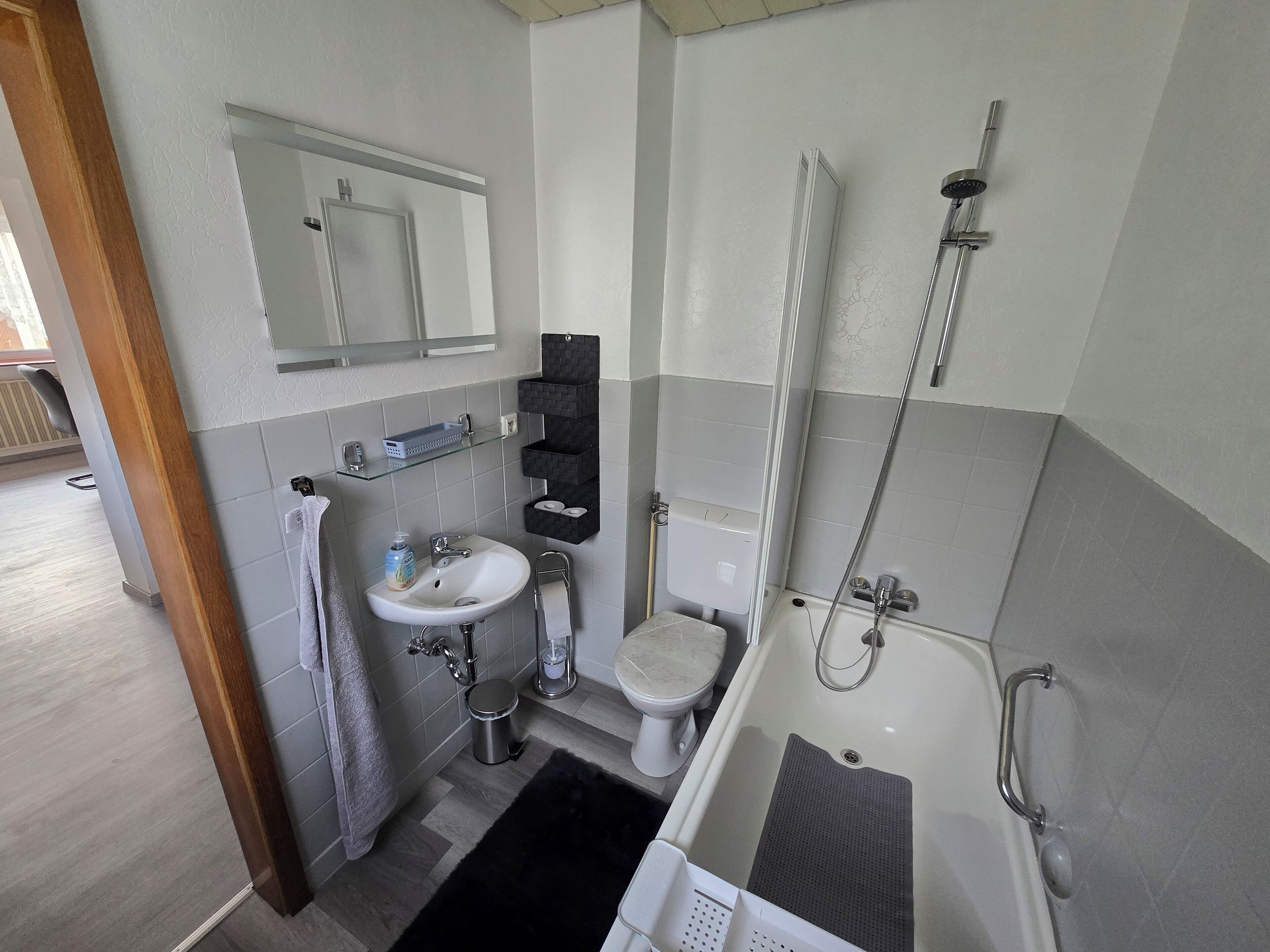 A modern bathroom with a sink, toilet, and shower. There is a mirror above the sink and a towel rack. The floor is tiled, and there is a black rug in the shower area.