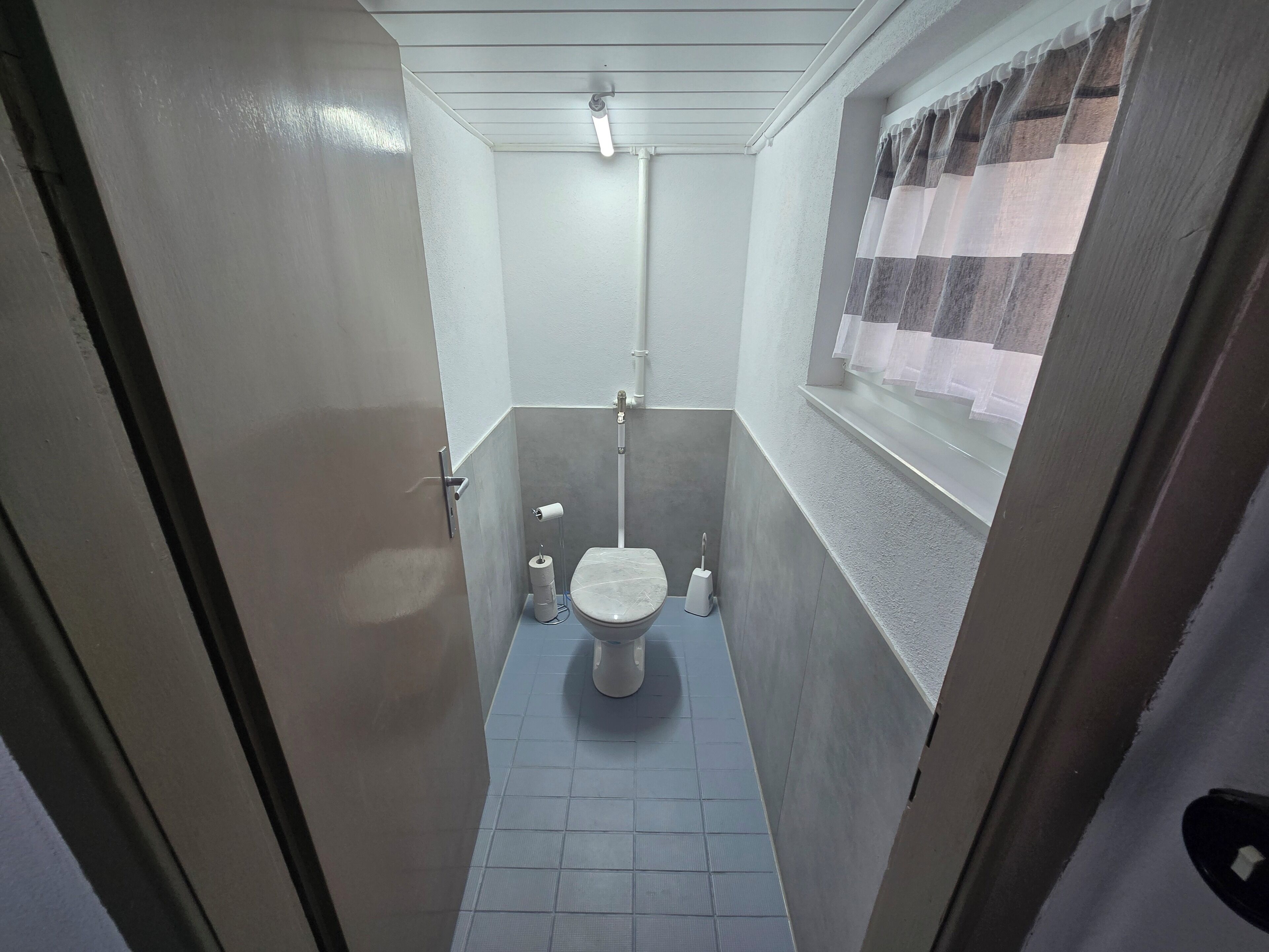 A small, narrow bathroom with a toilet, a shower, and a window with curtains.