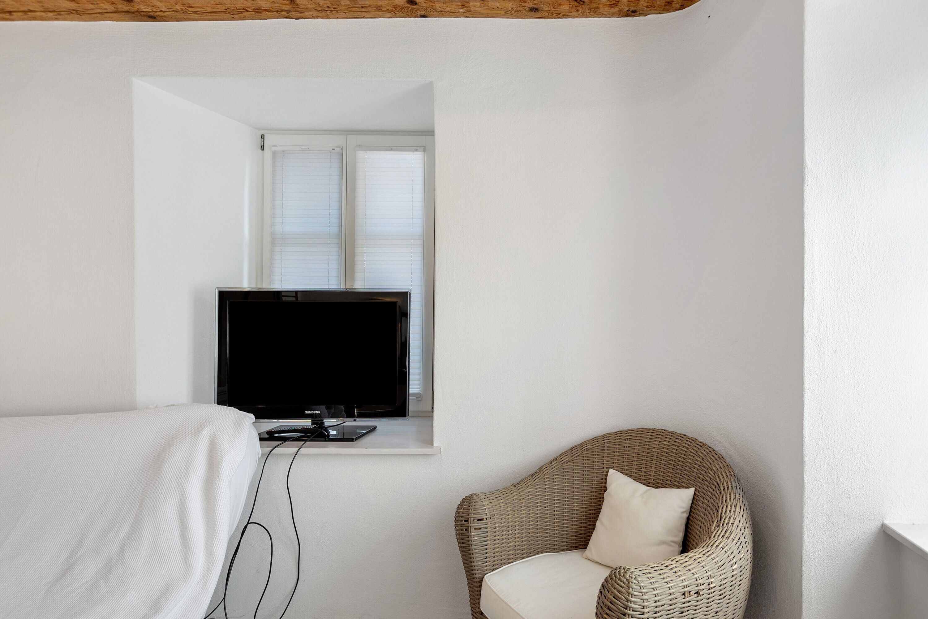A bedroom with a bed, a television on a shelf, a wicker chair, and a window with blinds.