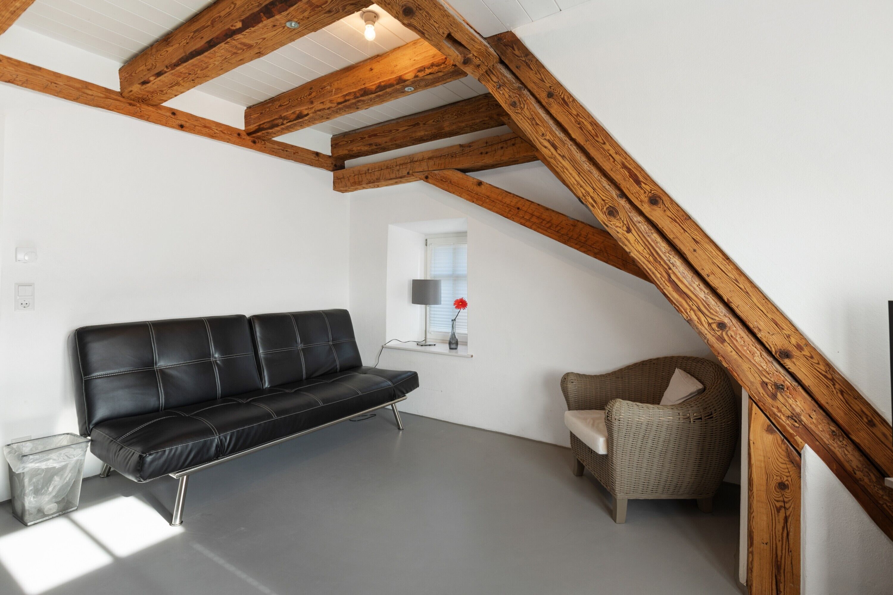 Une pièce avec un canapé en cuir noir, un fauteuil en osier, une petite fenêtre avec une lampe et des poutres apparentes.