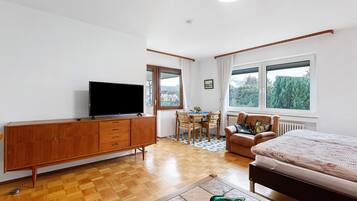 A bedroom with a bed, a TV on a wooden cabinet, a dining table with chairs, and large windows.