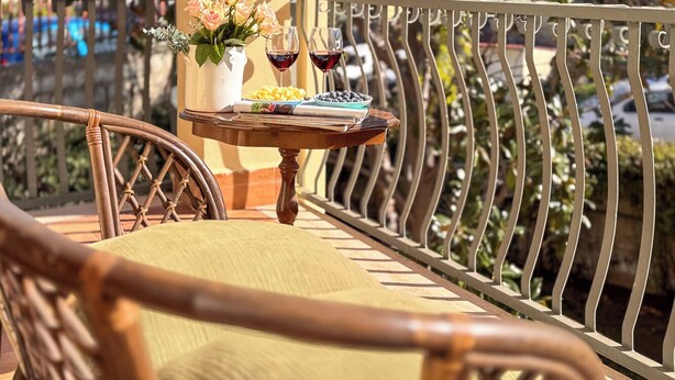 Un balcon avec des fauteuils en osier, une petite table sur laquelle est posé un vase de fleurs, deux verres de vin rouge et un saladier rempli de fruits.