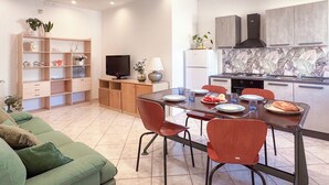 Un salon moderne avec un canapé vert, une table à manger avec quatre chaises, une cuisine équipée d’une cuisinière et d’un réfrigérateur, et un téléviseur posé sur un meuble en bois.