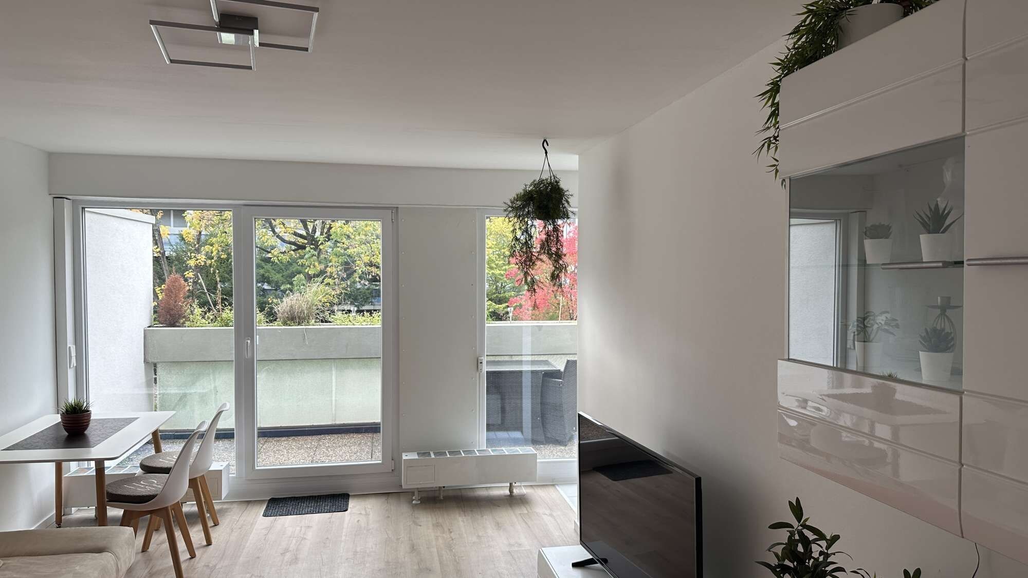 Una cocina moderna con horno empotrado, comedor con mesa y sillas, y puerta corrediza de vidrio que da a un balcón con vistas a árboles y un edificio.