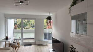 A modern kitchen with a built-in oven, a dining area with a table and chairs, and a sliding glass door leading to a balcony with a view of trees and a building. - Eastside Apartment Karlsruhe – stylish and peaceful living (Karlsruhe)