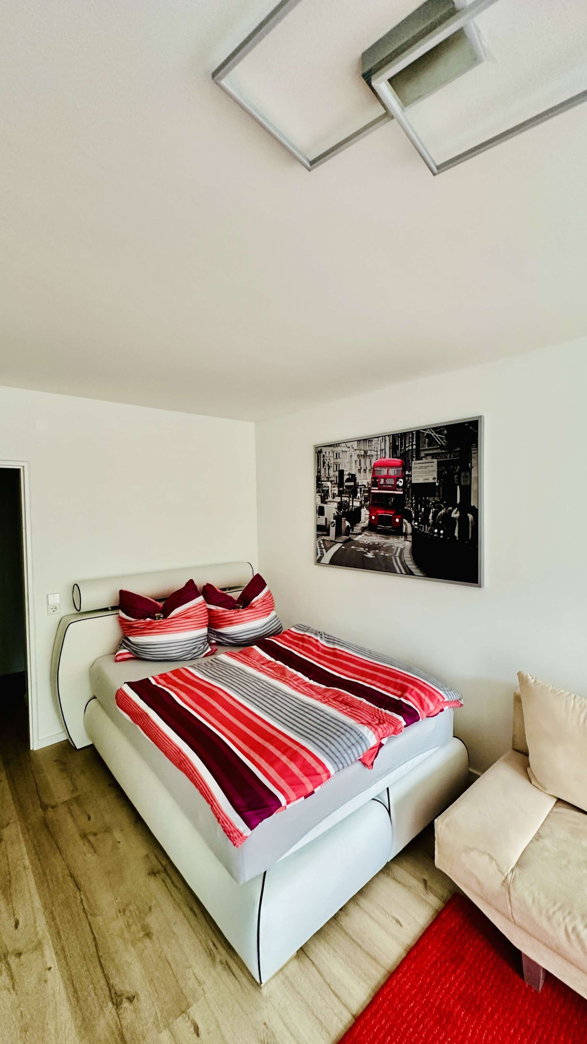 Ein Schlafzimmer mit einem Bett, einem Sessel, einem gerahmten Bild an der Wand und einer Deckenleuchte.