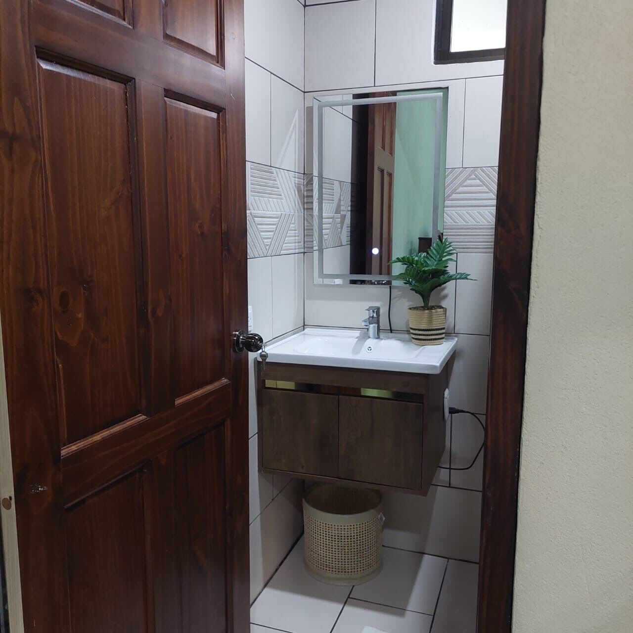 A bathroom with a wooden door, white tiled walls, a sink with a mirror, and a potted plant.