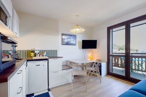 Eine kompakte Küche mit Kühlschrank, Herd und Mikrowelle. Es gibt einen Essbereich mit Tisch und Stühlen, einen an der Wand montierten Fernseher und einen Balkon mit Aussicht.