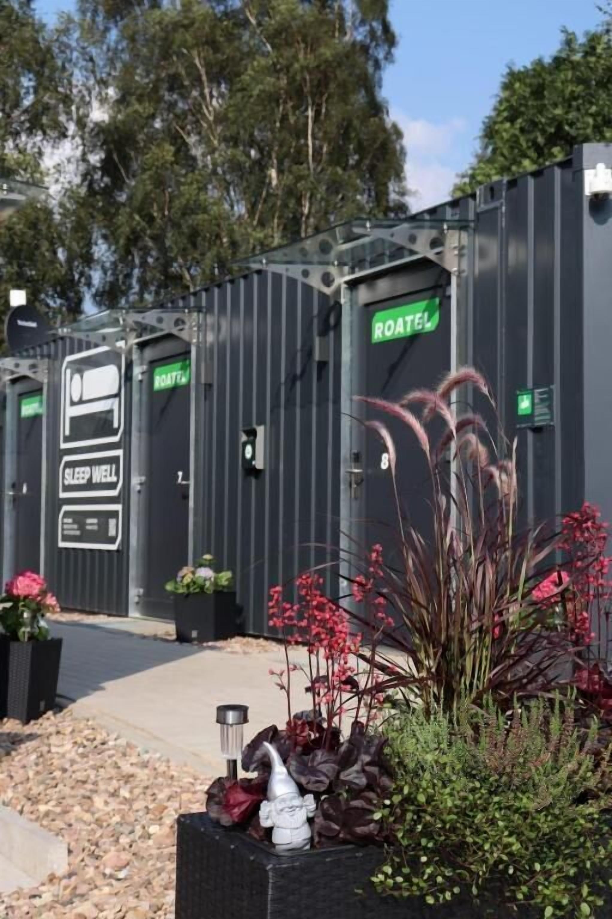 Portable restrooms with "SLEEP WELL" and "ROATEL" signs, surrounded by potted plants and a pathway.