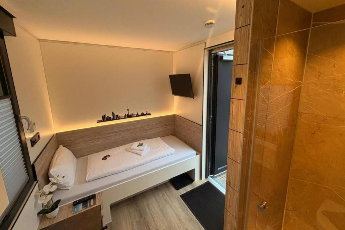 Ein modernes Hotelzimmer mit einem Bett, einem kleinen Nachttisch mit Pflanze, einem an der Wand montierten Fernseher und einem Holzschrank.
