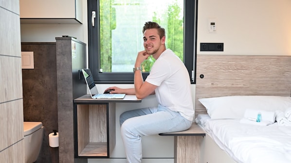 A modern room with a bed, a desk with a laptop, and a window with a view of greenery. - Roatel Holdorf A1 (Holdorf)
