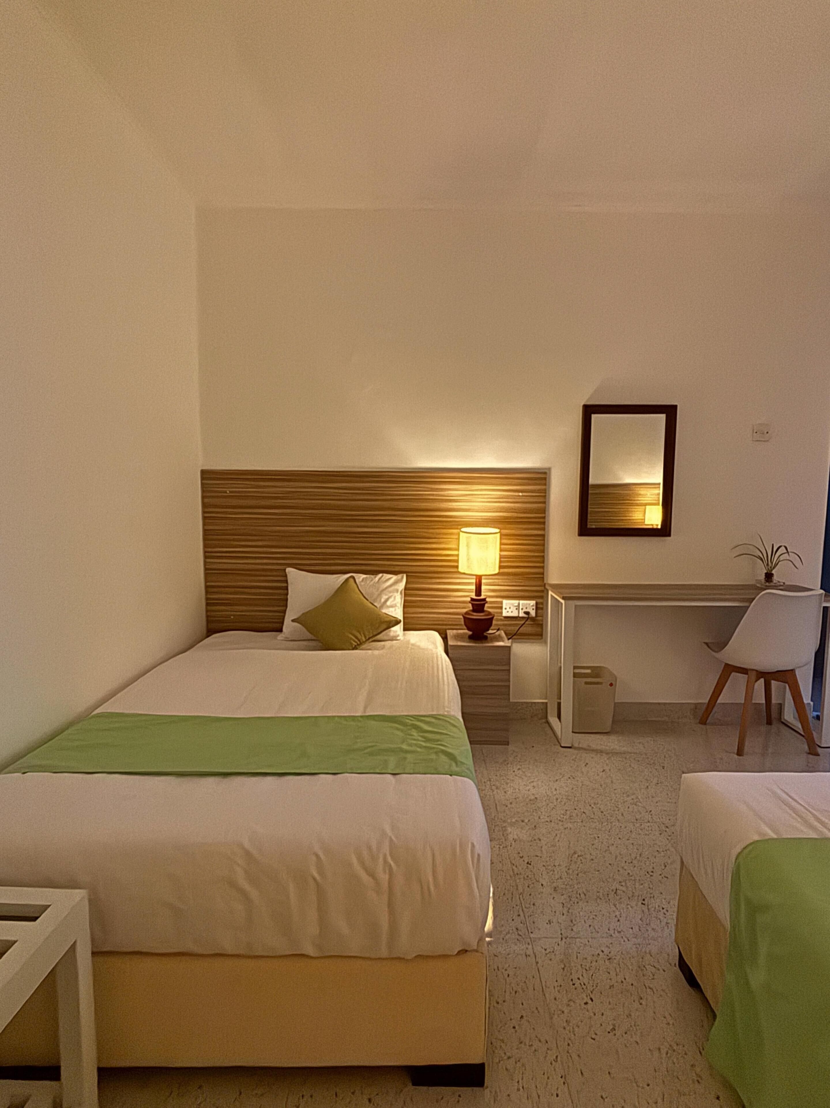 Hotellihuone, jossa on kaksi sänkyä, työpöytä, tuoli, lamppu, peili ja roskakori.
