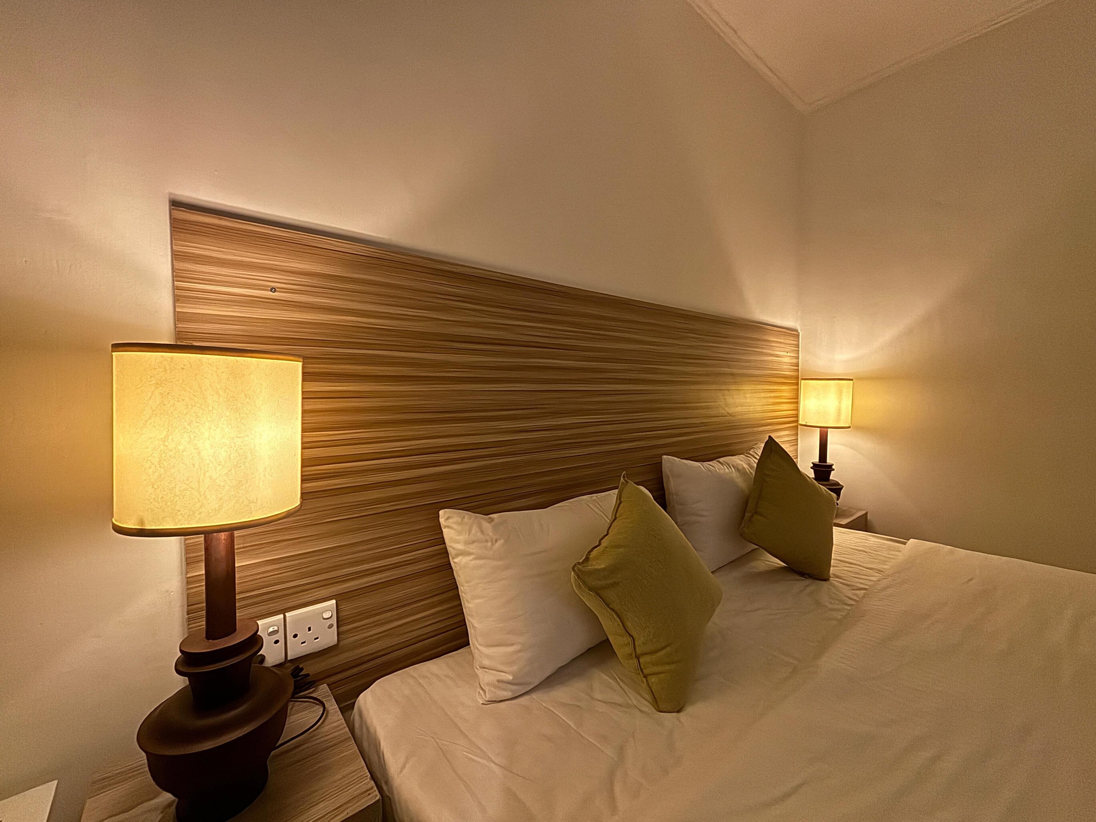 Habitació d'hotel amb un capçal de llit de fusta, un llit amb almoines blanques i verdes, i dues làmpades de nit.