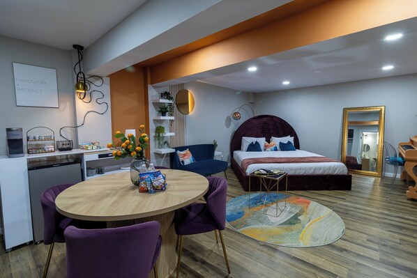 A modern hotel room with a wooden dining table, purple chairs, a bed with a headboard, a round rug, and a kitchenette.