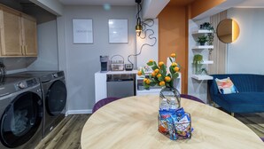 Une cuisine moderne équipée d’un lave-linge et d’un sèche-linge intégrés, un coin repas avec une table ronde et un espace de vie agrémenté d’un canapé bleu et de plantes décoratives.