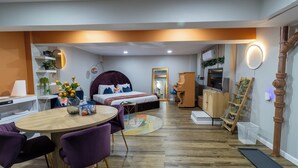 Une chambre d’hôtel moderne avec une table à manger en bois, des chaises violettes, un lit avec une tête de lit, une armoire en bois et une petite échelle en bois.