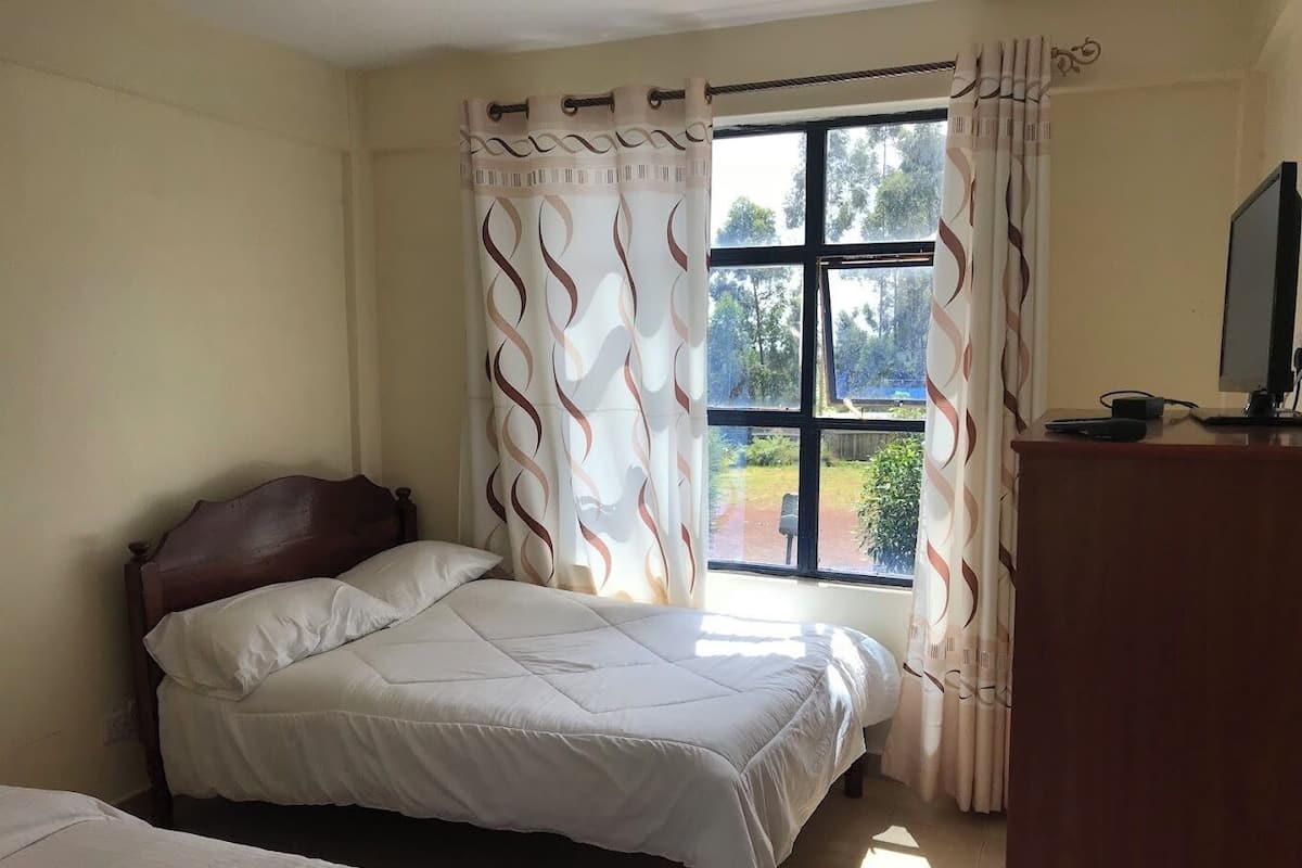 A bedroom with a bed, a wooden headboard, a desk with a computer, and a window with curtains.