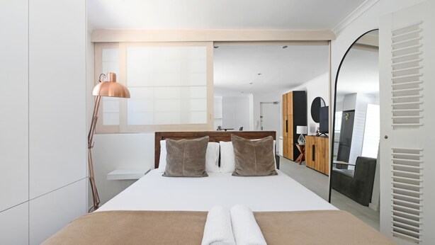 Une chambre d’hôtel moderne avec un grand lit, une tête de lit en bois, une grande fenêtre, un miroir et une armoire.