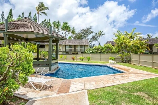 Um quintal com piscina, área de estar coberta e gramado cercado com plantas tropicais.