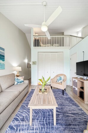 Una sala de estar con sofá, mesa de centro, sillón, mueble para televisor y ventilador de techo.