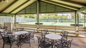 An outdoor patio area with tables and chairs, a tennis court visible through the background, and a covered roof structure. - Bright North Shore Condo With Loft, Golf Views, and Resort Amenities (Kahuku)
