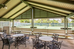 Un área de patio al aire libre con mesas y sillas, una cancha de tenis visible al fondo y una estructura de techo cubierta.