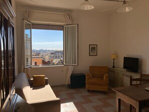A living room with a sofa, armchair, dining table, and a TV. A window offers a view of buildings and a clear sky. - Apartment with panoramic view - Holiday Home Giulia (Roma)