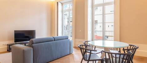 Una sala de estar moderna con un sofá gris, una mesa de comedor de vidrio con sillas, un televisor de pantalla plana y ventanas amplias con vista a la ciudad.