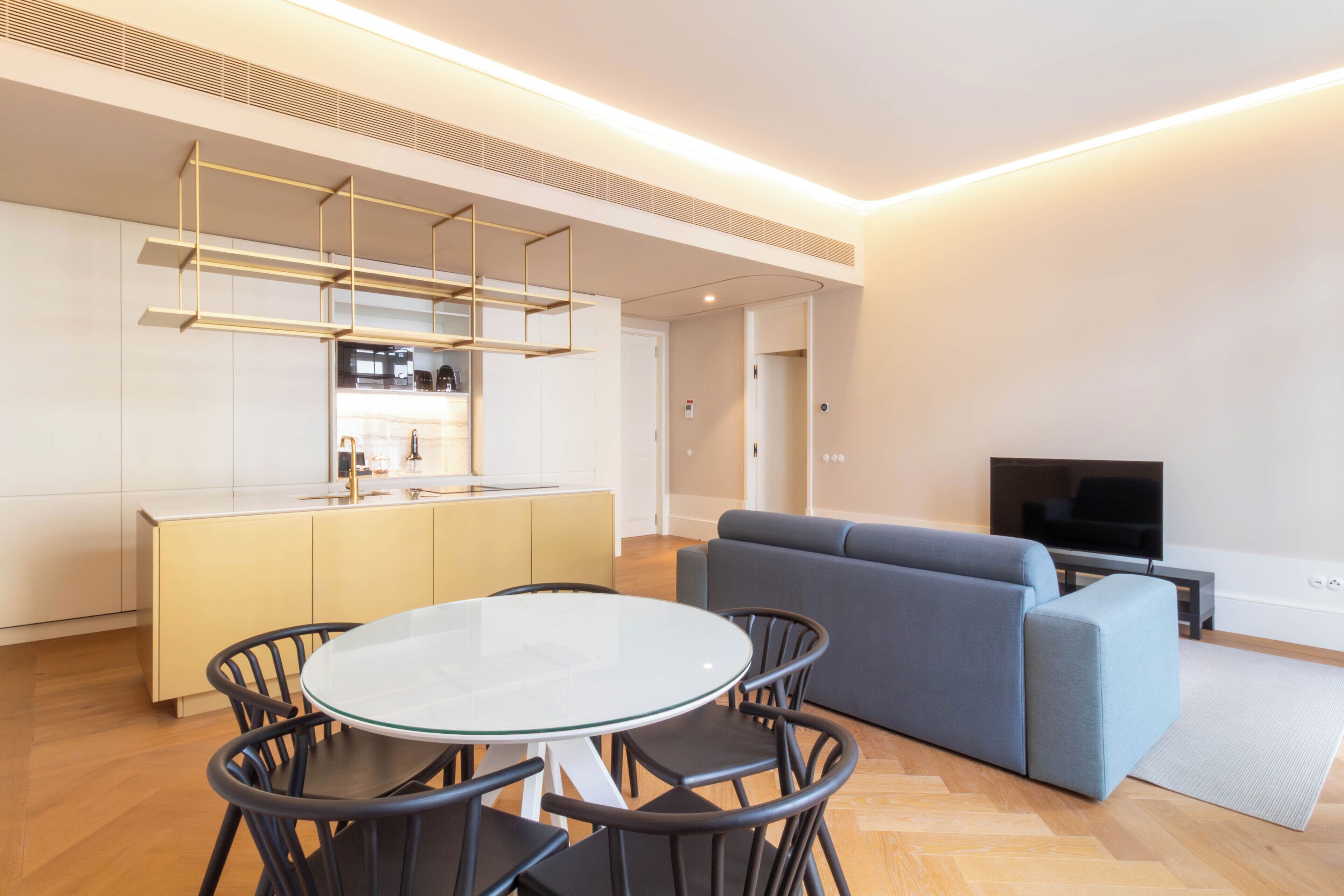 A modern living room with a grey sofa, a round glass table with chairs, and a kitchen area with white cabinets and a built-in TV.