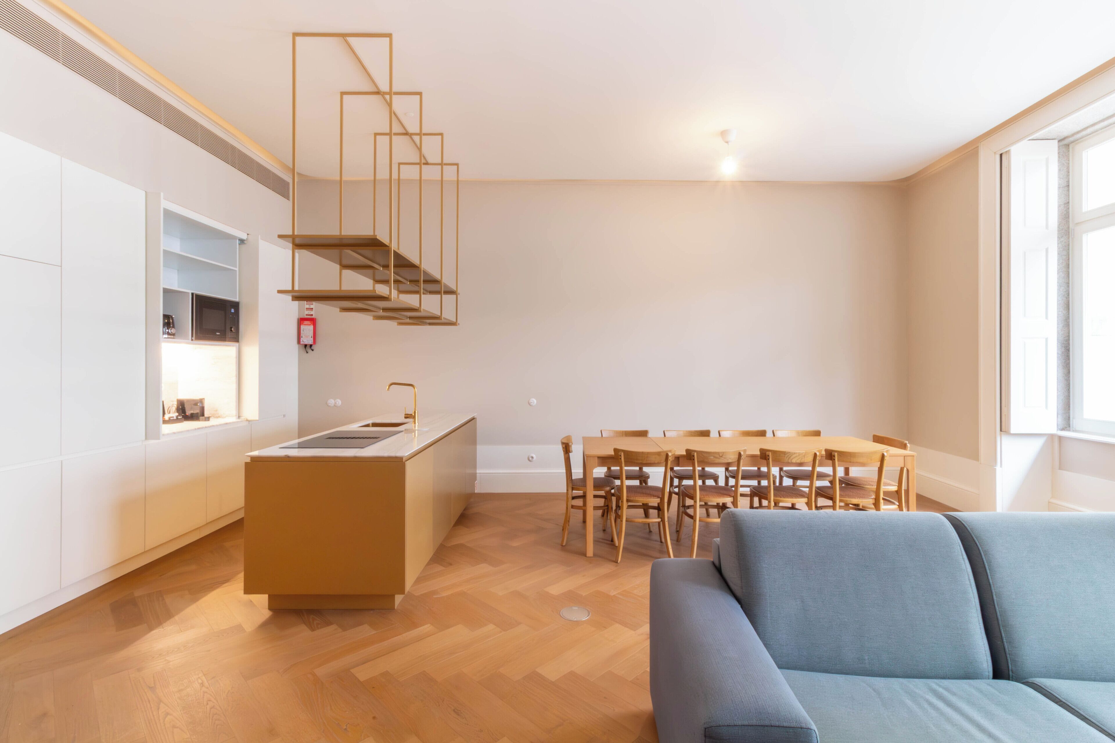 A modern kitchen with a central island, wooden dining table with chairs, and a sofa in a bright living area with large windows.