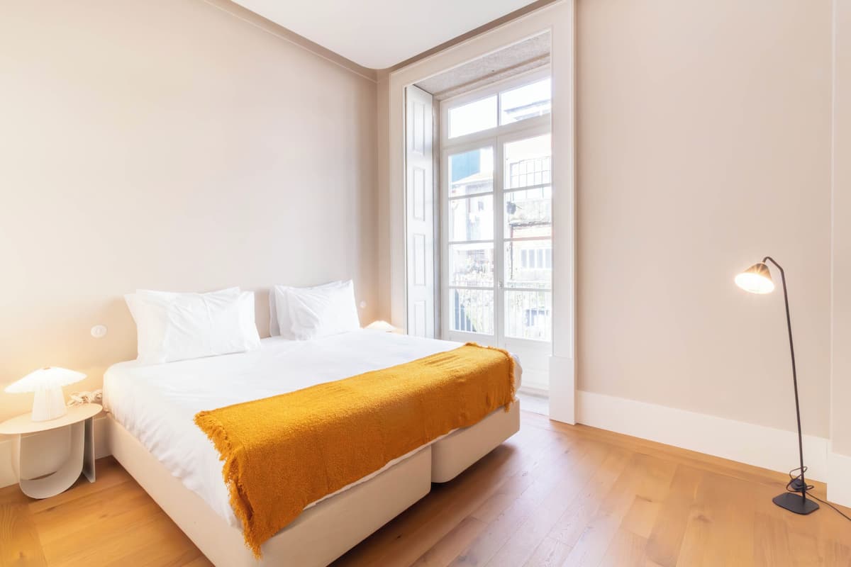 Une chambre à coucher avec un grand lit, une table de chevet, une lampe sur pied et une fenêtre donnant sur des immeubles.