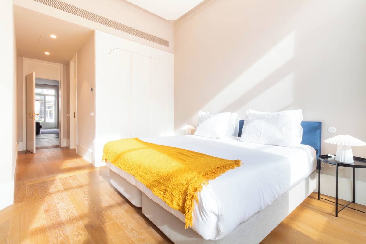 Une chambre moderne avec un grand lit, un plaid jaune, une table de chevet avec une lampe et une vue sur un couloir.