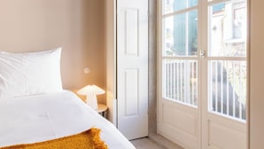 A bedroom with a bed, a nightstand, a lamp, and a door leading to a balcony with a view of buildings. - Entreparedes House (Porto)