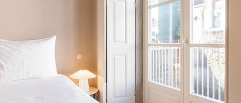 A bedroom with a bed, a nightstand, a lamp, and a door leading to a balcony with a view of buildings.