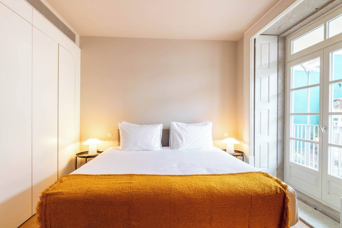 Une chambre d’hôtel avec un lit, deux tables de chevet avec des lampes, une fenêtre donnant sur un paysage agréable et une porte.