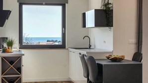 A modern kitchen with a large window, white cabinets, a black countertop, and a dining area with a chair and a bowl of fruit. - Apartment Citylight | Belvedere Holidays – Sea Views & Modern Comfort (Novigrad)