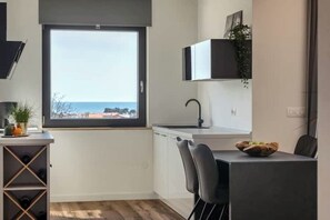 Eine moderne Küche mit großem Fenster, weißen Schränken, einer schwarzen Arbeitsplatte und einem Essbereich mit einem Stuhl und einer Fruchtschale.