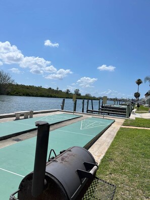 Ein sonniger Außenbereich mit Shuffleboard-Tischen, einem Grill und Blick auf das Wasser.