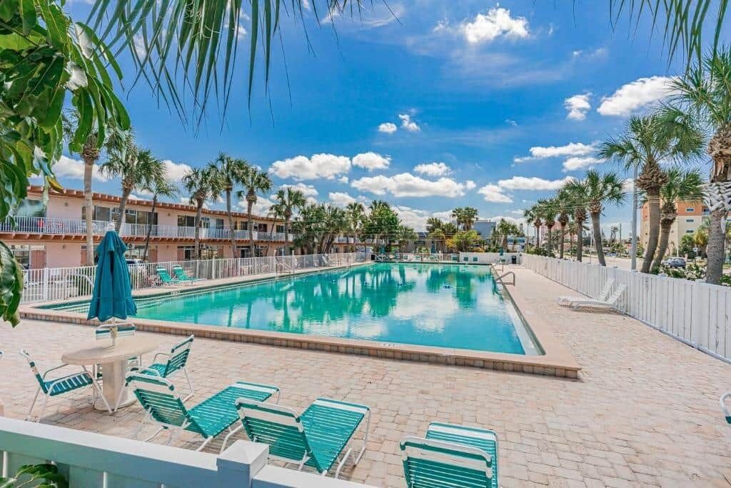 A swimming pool with lounge chairs and umbrellas, surrounded by palm trees and a building with a white fence.
